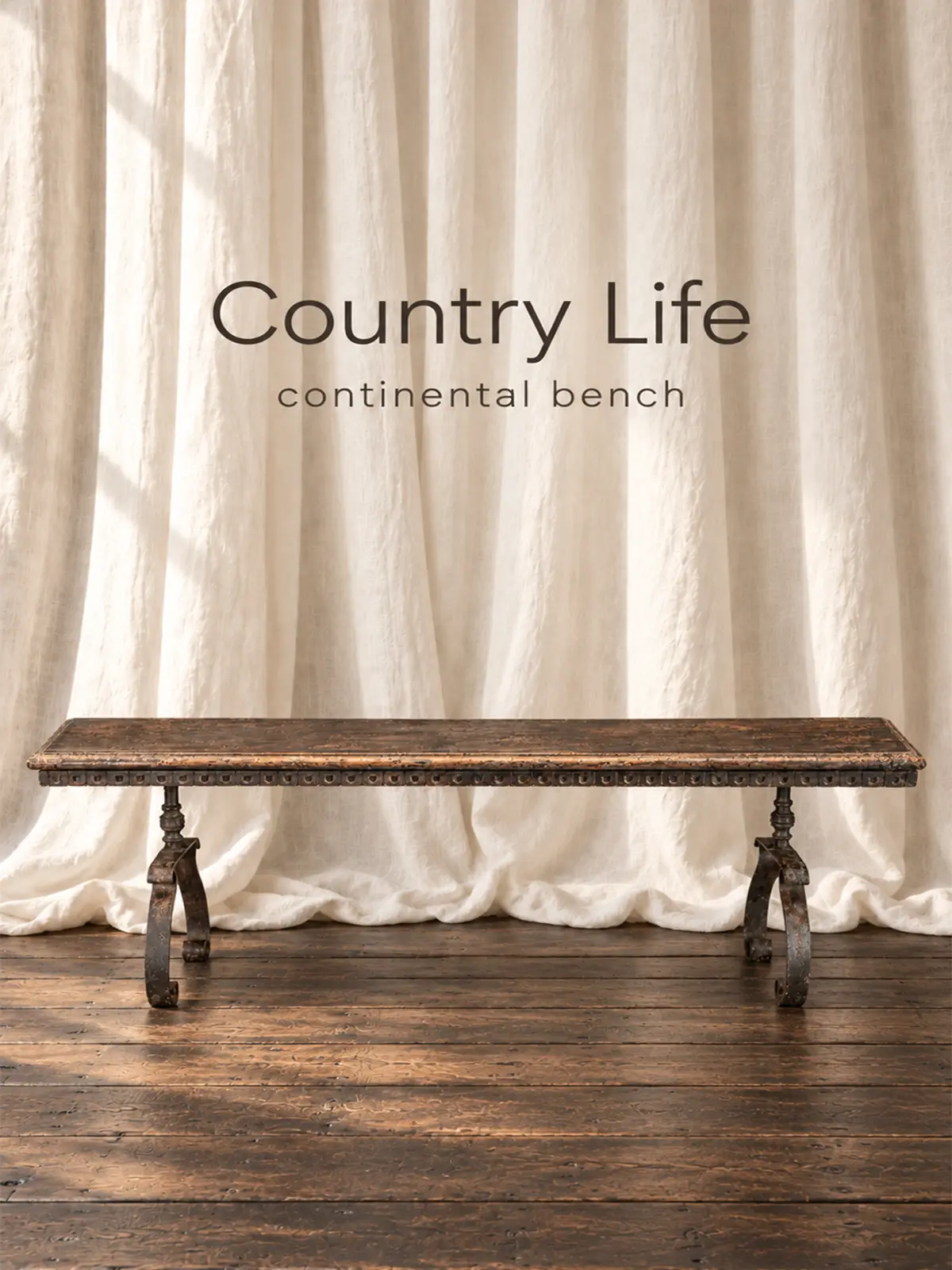 Antique Continental bench with a timeworn wooden seat and curved iron legs, displayed before ivory linen curtains on reclaimed wood flooring.